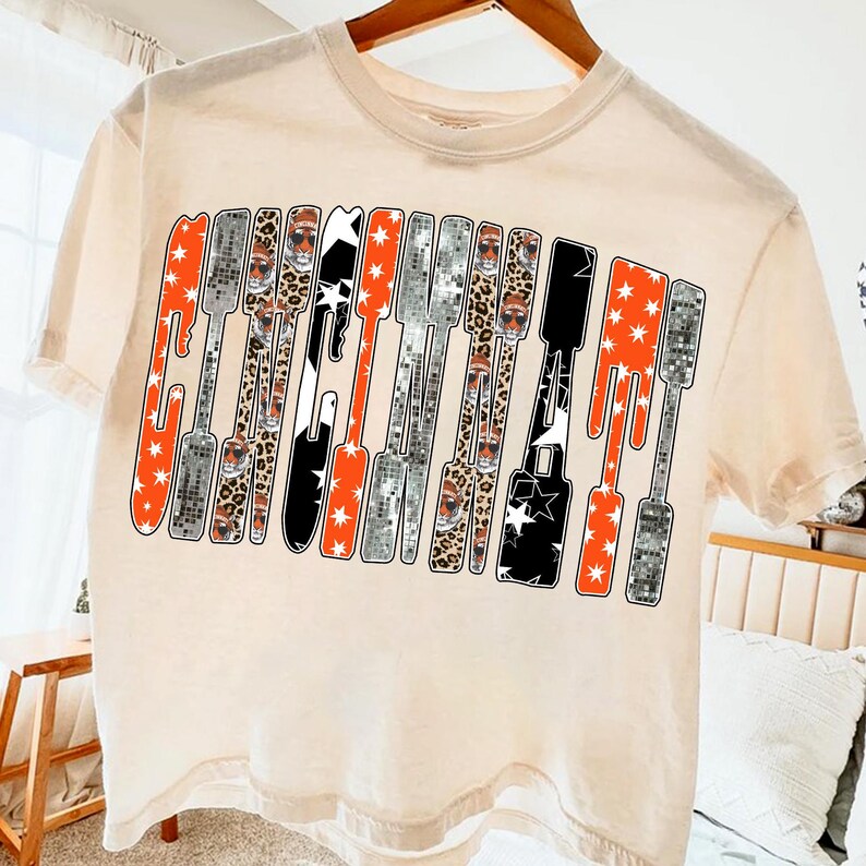 Puede incluir: Camiseta color crema con la palabra "Guitar" en un dise&ntilde;o estilizado. Las letras est&aacute;n compuestas por varios patrones, incluyendo naranja con estrellas blancas, estampado de leopardo, negro con estrellas blancas y patrones de bola de discoteca plateada.