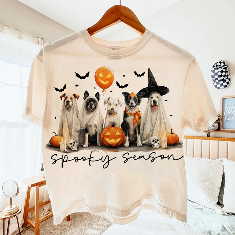 May include: Cream-colored t-shirt featuring a Halloween design. The graphic includes five dogs dressed in costumes, pumpkins, bats, and the text "Spooky Season". A festive and fun design for the fall.