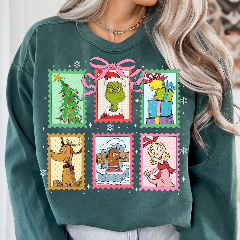 May include: Dark green sweatshirt featuring a colorful Christmas-themed design. The design includes six postage stamp-style images with characters and elements from a holiday story. The images include a Christmas tree, a green character, wrapped gifts, a reindeer, a snowy scene, and a girl.