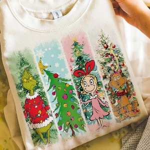 Op de afbeelding: Cr&egrave;mekleurige sweatshirt met een kleurrijke afbeelding van personages en kerstbomen. Het ontwerp bevat de Grinch, Cindy Lou Who, Max en versierde bomen in verticale panelen. De Grinch draagt een rode trui.