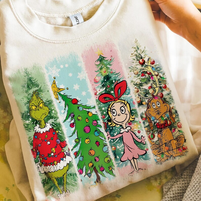 Op de afbeelding: Cr&egrave;mekleurige sweatshirt met een kleurrijke afbeelding van personages en kerstbomen. Het ontwerp bevat de Grinch, Cindy Lou Who, Max en versierde bomen in verticale panelen. De Grinch draagt een rode trui.
