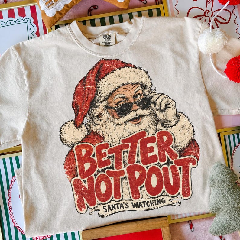 May include: A cream-colored short-sleeve t-shirt featuring a graphic of Santa Claus wearing sunglasses and a red hat. The text "BETTER NOT POUT" is printed in large, distressed red letters below, with "SANTA'S WATCHING" in smaller black text.