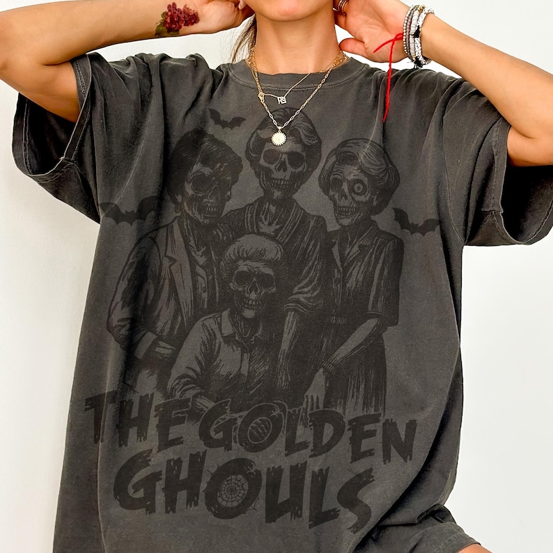 Puede incluir: Camiseta gris oscuro con un gr&aacute;fico de cuatro figuras esquel&eacute;ticas y el texto "THE GOLDEN GHOULS". El dise&ntilde;o incluye murci&eacute;lagos. La camiseta tiene un aspecto vintage y desgastado.