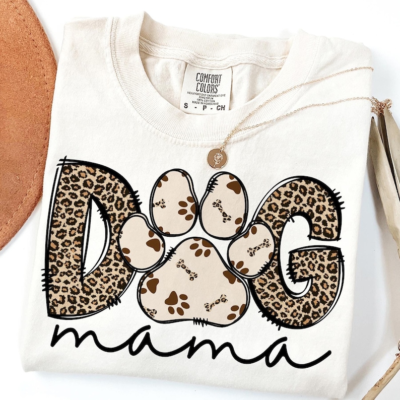 Pu&ograve; includere: Una maglietta bianca con un design a stampa leopardata marrone e nera che dice "Dog mama". Il "Dog" &egrave; composto da un'impronta di zampa e le lettere "D" e "G" sono in stampa leopardata.