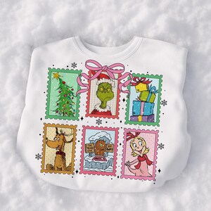May include: White sweatshirt featuring six postage stamp-style images of characters and scenes from a holiday story. The images include a Christmas tree, a green character, wrapped gifts, a dog, a door with text, and a girl. A pink bow is at the top.