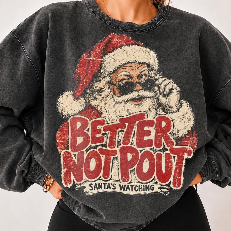 May include: A charcoal gray crewneck sweatshirt featuring a distressed graphic of Santa Claus wearing sunglasses. The text reads "BETTER NOT POUT" in large red letters, with "SANTA'S WATCHING" printed in smaller white letters underneath the graphic.