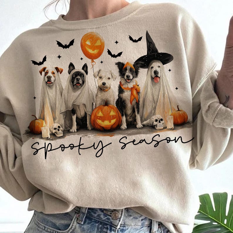 May include: Beige sweatshirt featuring a Halloween design. The graphic includes five dogs dressed in costumes, pumpkins, bats, and the phrase "Spooky Season". A pumpkin balloon is also present in the design.