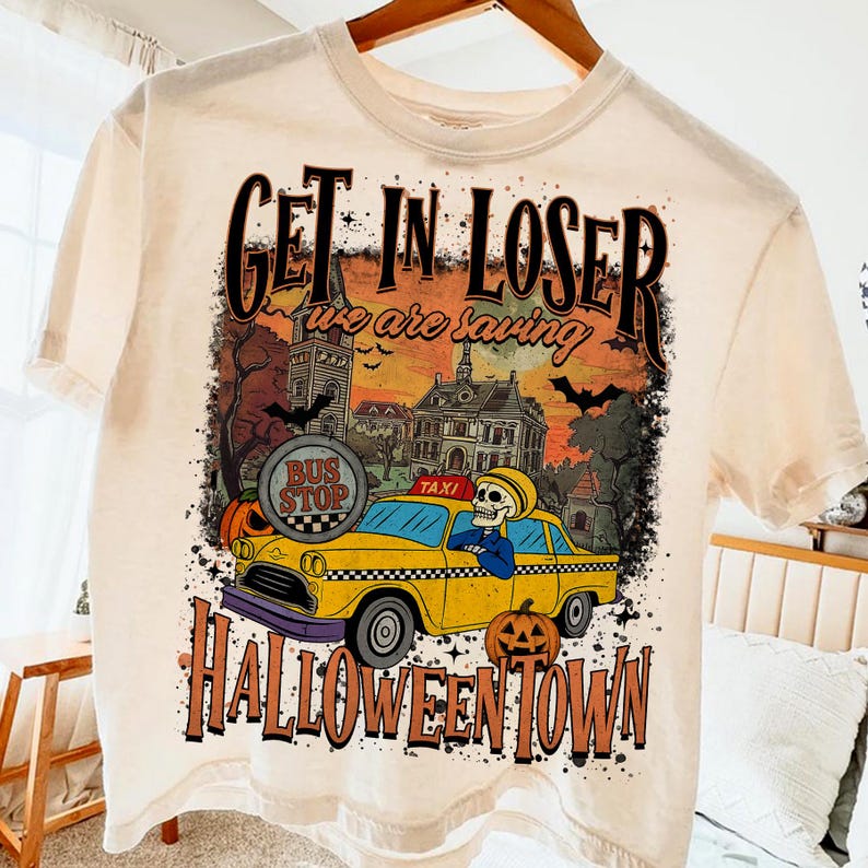 May include: Cream-colored t-shirt with a Halloween-themed graphic. The design features a yellow taxi with a skeleton driver, pumpkins, and the text "Get in Loser we are saving Halloween Town."