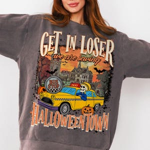 May include: Dark gray sweatshirt with a Halloween-themed graphic. The design features a yellow taxi with a skeleton driver, pumpkins, bats, and the text "GET IN LOSER we are saving HALLOWEENTOWN". The graphic has a vintage, distressed look.