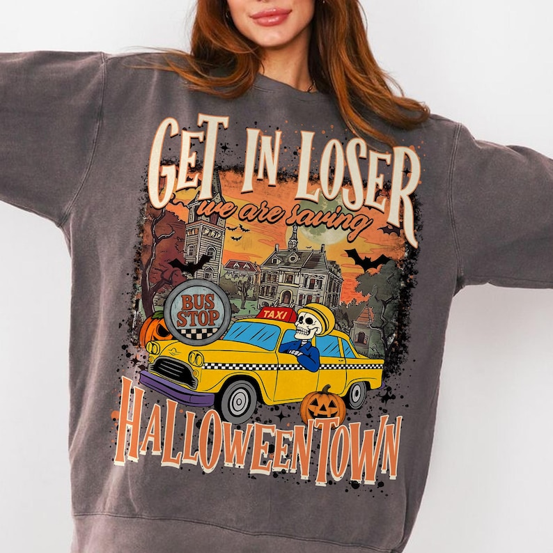 May include: Dark gray sweatshirt with a Halloween-themed graphic. The design features a yellow taxi with a skeleton driver, pumpkins, bats, and the text "GET IN LOSER we are saving HALLOWEENTOWN". The graphic has a vintage, distressed look.
