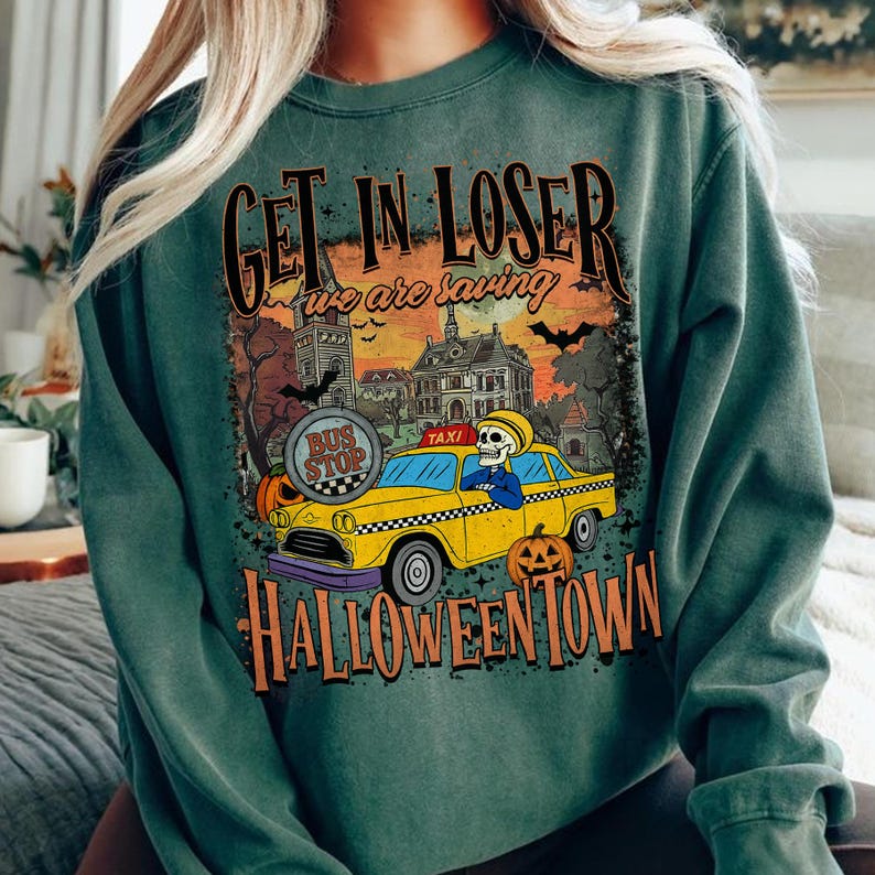 May include: Green sweatshirt with a Halloween-themed graphic. The design features a yellow taxi with a skeleton driver, pumpkins, and the text "Get in Loser, we are saving Halloween Town."