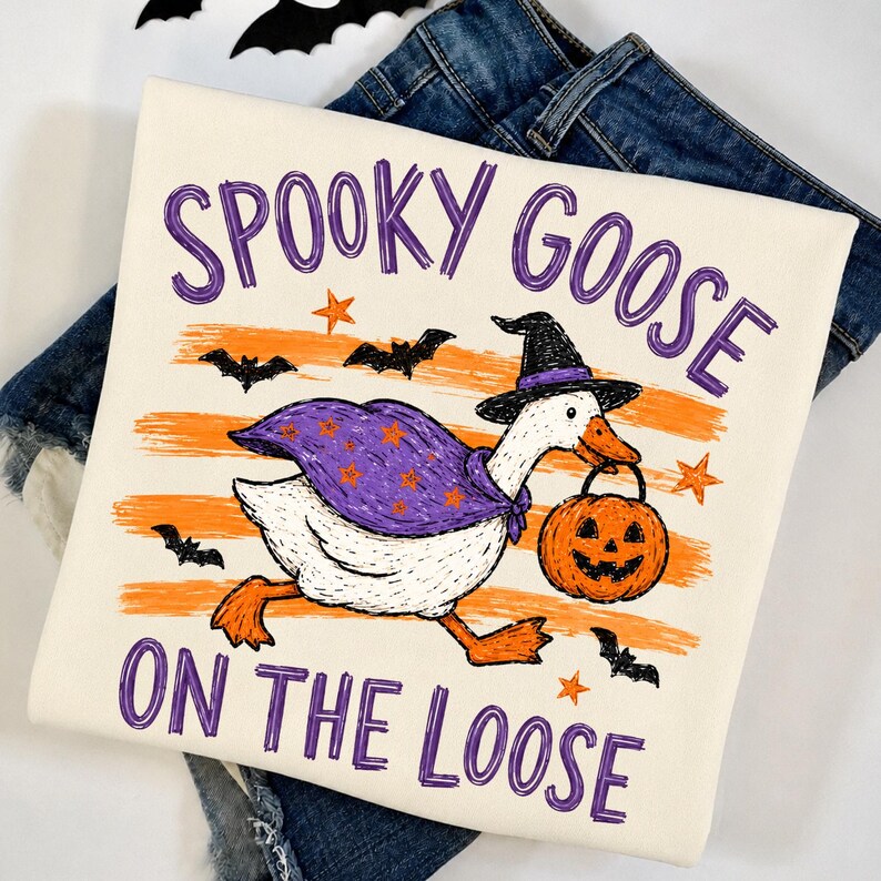 May include: A cream-colored t-shirt featuring a graphic of a white goose wearing a purple star-patterned cape and a black witch hat. The goose carries an orange jack-o'-lantern. The text reads SPOOKY GOOSE ON THE LOOSE in purple letters.
