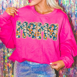 May include: A pink sweatshirt with the word "MAMA" in large, white letters outlined in gold. The letters are covered in a floral pattern.