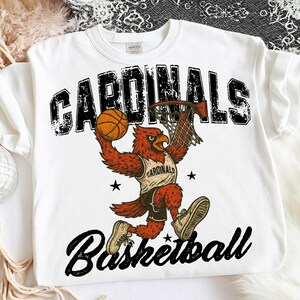 May include: White sweatshirt featuring a red cardinal bird dunking a basketball. The word "CARDINALS" is printed above the bird in a distressed black font, with "Basketball" below. The bird wears a jersey that says "CARDINALS".