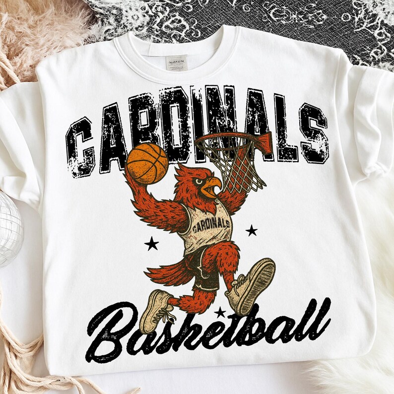 May include: White sweatshirt featuring a red cardinal bird dunking a basketball. The word "CARDINALS" is printed above the bird in a distressed black font, with "Basketball" below. The bird wears a jersey that says "CARDINALS".