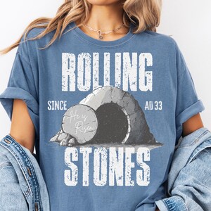 May include: A blue t-shirt with the words "ROLLING STONES" in large white letters. The shirt features a graphic of a tomb with a stone rolled away, and the text "He is Risen" on the stone. The shirt also has the text "SINCE AD 33".