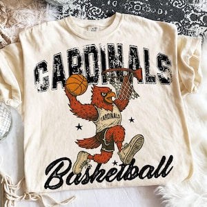 May include: Cream-colored t-shirt featuring a basketball-playing cardinal mascot. The design includes the word "CARDINALS" above the mascot and "Basketball" below. The cardinal is depicted dunking a basketball into a hoop.