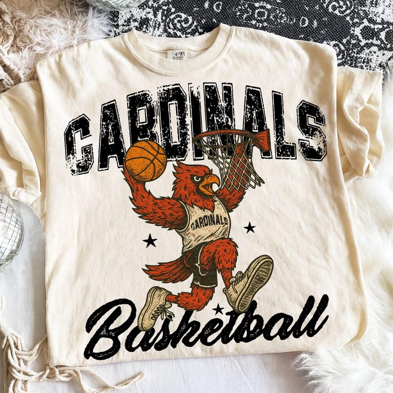 May include: Cream-colored t-shirt featuring a basketball-playing cardinal mascot. The design includes the word "CARDINALS" above the mascot and "Basketball" below. The cardinal is depicted dunking a basketball into a hoop.