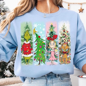 Op de afbeelding: Lichtblauw sweatshirt met een kerstontwerp. De afbeelding toont de Grinch, een versierde kerstboom, Cindy Lou Who en Max de hond, gerangschikt in verticale panelen. Feestelijk thema.