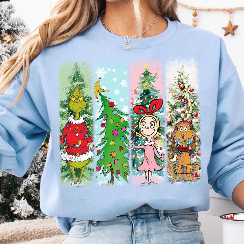 Op de afbeelding: Lichtblauw sweatshirt met een kerstontwerp. De afbeelding toont de Grinch, een versierde kerstboom, Cindy Lou Who en Max de hond, gerangschikt in verticale panelen. Feestelijk thema.