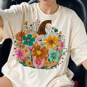May include: Cream-colored t-shirt featuring a colorful floral pumpkin design. The pumpkin is filled with orange, yellow, pink, and teal flowers, with a brown stem. A casual, oversized fit, perfect for fall.