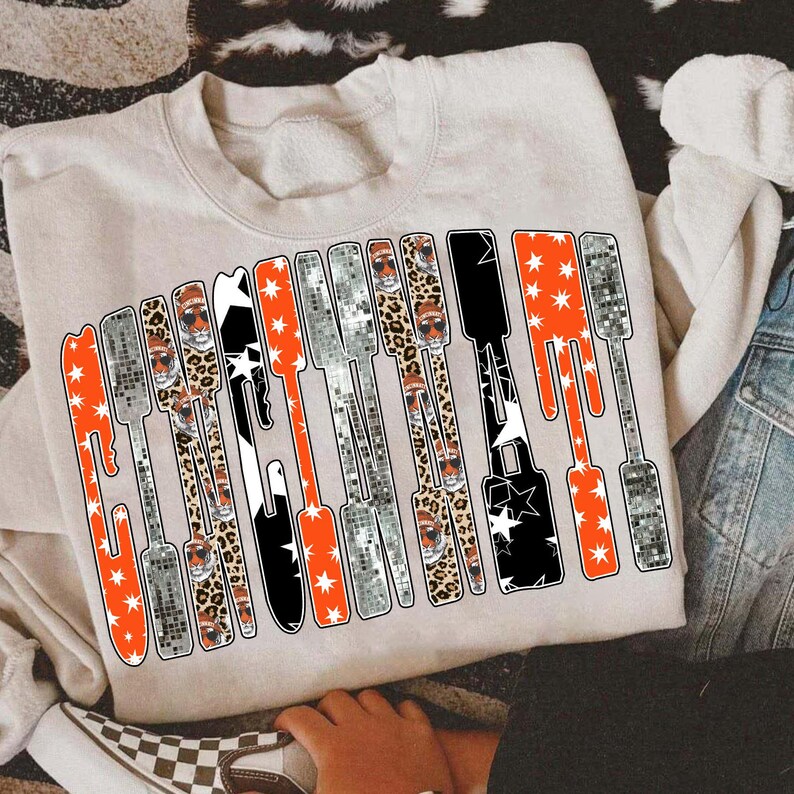 Puede incluir: Sudadera beige con la palabra "Gameday" en un dise&ntilde;o estilizado. Las letras incorporan patrones como estrellas naranjas, efectos de bola de discoteca, estampado de leopardo y patrones de estrellas en blanco y negro. Un estilo casual y c&oacute;modo.