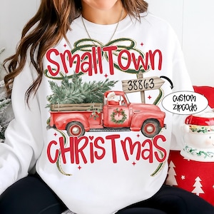 May include: White sweatshirt with a red graphic of a vintage red truck carrying a Christmas tree. The text "Small Town Christmas" is above and below the truck. A sign reads "38863" and a custom zipcode option is shown.