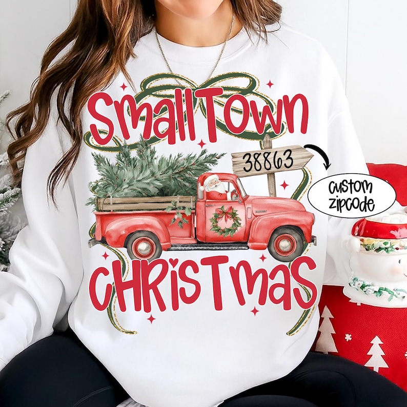 May include: White sweatshirt with a red graphic of a vintage red truck carrying a Christmas tree. The text "Small Town Christmas" is above and below the truck. A sign reads "38863" and a custom zipcode option is shown.