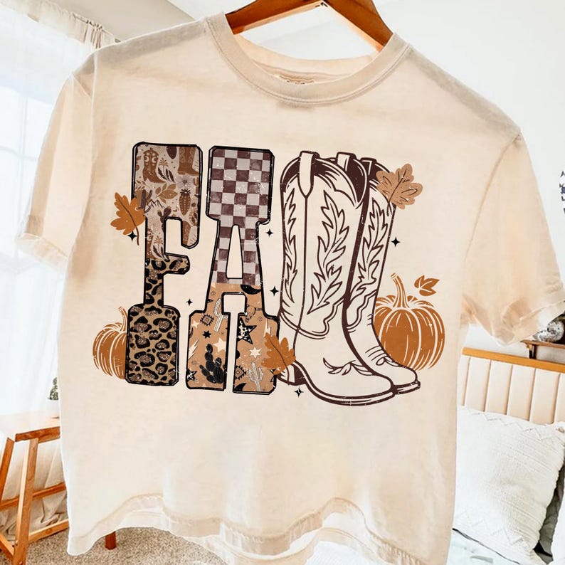 K&ouml;nnte beinhalten: Cremefarbenes T-Shirt mit dem Wort "FALL" in einem dekorativen Design. Die Buchstaben zeigen Muster wie Leopardenmuster, ein Schachbrettmuster und Cowboy-Stiefel. Braune K&uuml;rbisse und Bl&auml;tter akzentuieren das Design und evozieren ein Herbstthema.