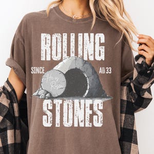 May include: Brown t-shirt with the text "ROLLING STONES" in white, along with an illustration of a tomb and the words "He is Risen." The shirt also includes the text "SINCE AD 33."