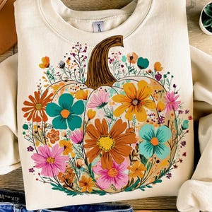 May include: Cream-colored sweatshirt featuring a large pumpkin design filled with colorful flowers. The pumpkin has a brown stem and is surrounded by orange, teal, pink, and yellow floral accents. The sweatshirt is folded, with a portion of the sleeve visible.