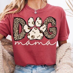 Pu&ograve; includere: Una maglietta rossa con una grafica di un'impronta di zampa composta dalle lettere "DOG" in un motivo leopardato. Il testo "mama" &egrave; scritto sotto l'impronta di zampa.