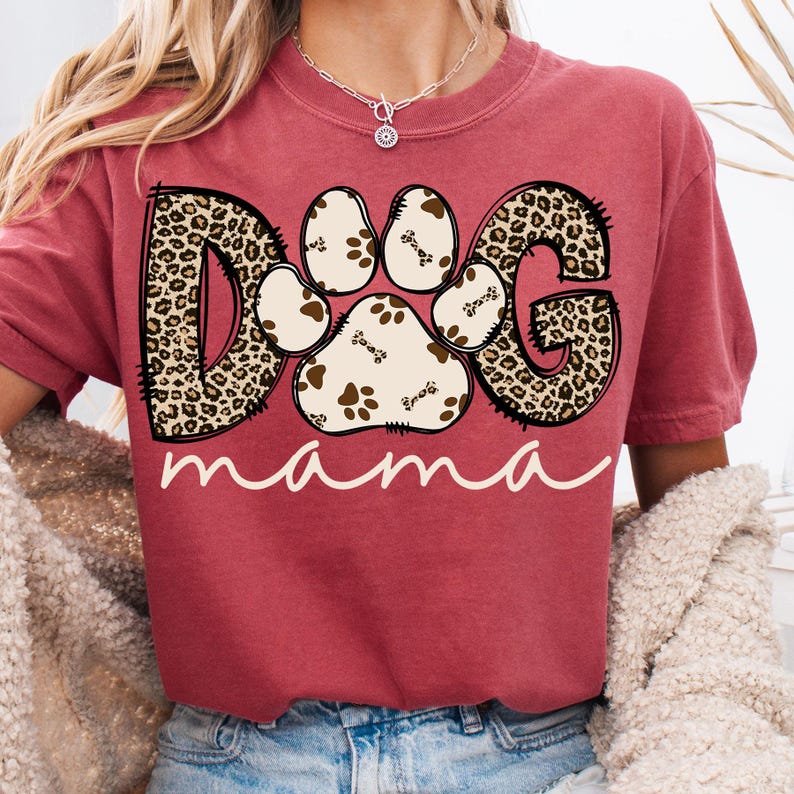 Pu&ograve; includere: Una maglietta rossa con una grafica di un'impronta di zampa composta dalle lettere "DOG" in un motivo leopardato. Il testo "mama" &egrave; scritto sotto l'impronta di zampa.