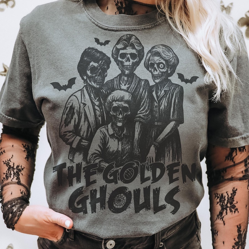 Puede incluir: Camiseta gris con un gr&aacute;fico negro de cuatro figuras esquel&eacute;ticas y murci&eacute;lagos. El texto "THE GOLDEN GHOULS" est&aacute; impreso debajo de las figuras en una fuente desgastada. La camiseta tiene mangas cortas y cuello redondo.