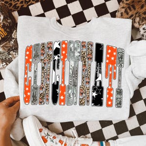 Puede incluir: Sudadera gris claro con un dise&ntilde;o gr&aacute;fico en naranja, negro, plateado y estampado de leopardo. El dise&ntilde;o deletrea una palabra en una fuente vertical y alargada con acentos de estrellas. Se ven zapatillas blancas.