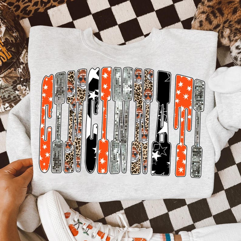 Puede incluir: Sudadera gris claro con un dise&ntilde;o gr&aacute;fico en naranja, negro, plateado y estampado de leopardo. El dise&ntilde;o deletrea una palabra en una fuente vertical y alargada con acentos de estrellas. Se ven zapatillas blancas.
