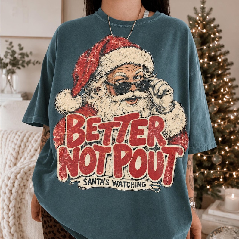 May include: A dark teal short-sleeve t-shirt featuring a graphic of Santa Claus wearing sunglasses. The text reads "BETTER NOT POUT" in large red letters with "SANTA'S WATCHING" underneath. The design has a distressed, vintage-inspired texture.