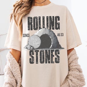 May include: A beige t-shirt with the words "ROLLING STONES" in a distressed font. The graphic depicts a tomb with the text "He is Risen" and the dates "SINCE AD 33". The shirt has a relaxed fit.