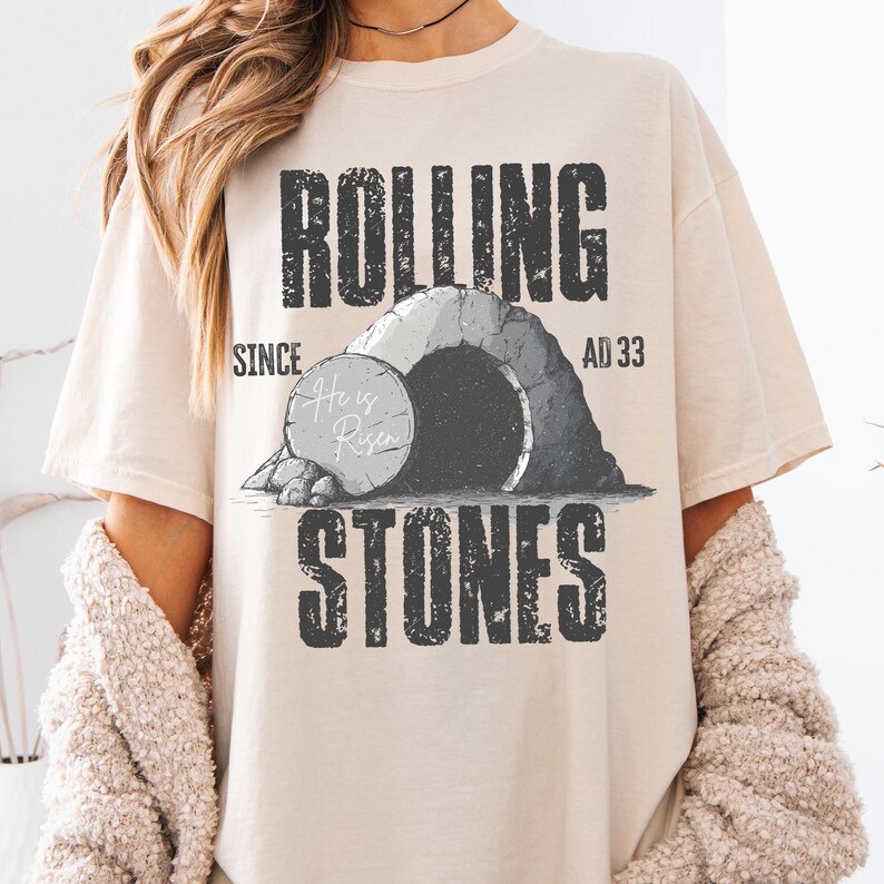 May include: A beige t-shirt with the words "ROLLING STONES" in a distressed font. The graphic depicts a tomb with the text "He is Risen" and the dates "SINCE AD 33". The shirt has a relaxed fit.