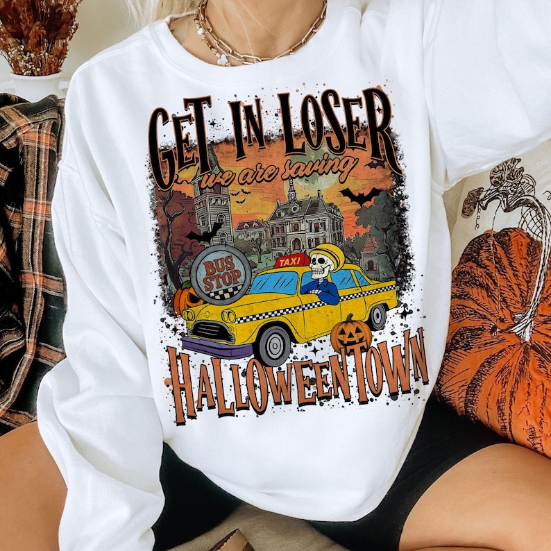 May include: White long-sleeve sweatshirt with a Halloween-themed graphic. The design features a yellow taxi with a skeleton driver, pumpkins, and the text "GET IN LOSER we are saving HALLOWEENTOWN".