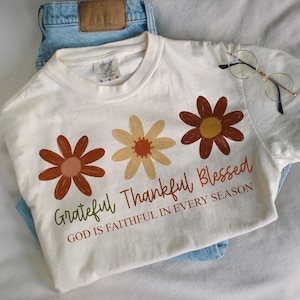 May include: Cream-colored t-shirt with three floral designs in shades of brown and yellow. The shirt features the text "Grateful Thankful Blessed" and "God is Faithful in Every Season" in a script font. The shirt is laid on top of blue jeans.