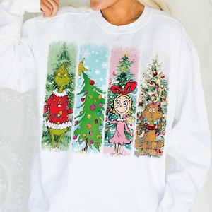 Op de afbeelding: Witte sweatshirt met lange mouwen met een kleurrijke afbeelding van personages en kerstbomen. Het ontwerp bevat de Grinch, een versierde boom, Cindy Lou Who en Max de hond, allemaal in een feestelijk thema.