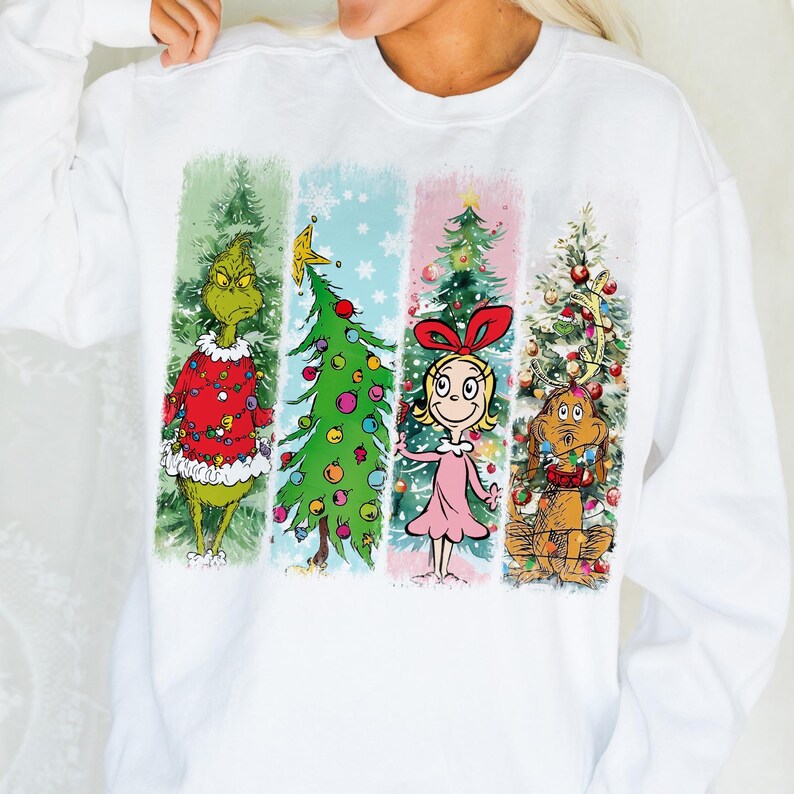 Op de afbeelding: Witte sweatshirt met lange mouwen met een kleurrijke afbeelding van personages en kerstbomen. Het ontwerp bevat de Grinch, een versierde boom, Cindy Lou Who en Max de hond, allemaal in een feestelijk thema.