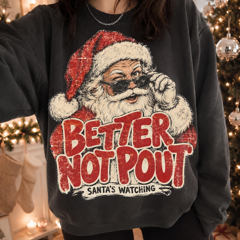 May include: A charcoal gray crewneck sweatshirt featuring a graphic of Santa Claus wearing sunglasses and a red hat. The text reads "BETTER NOT POUT" in large red letters, with "SANTA'S WATCHING" printed in smaller black text underneath the graphic.