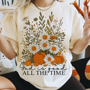 May include: Cream-coloured t-shirt with a floral and pumpkin design. The graphic includes white daisies, orange pumpkins, and the text "God is good all the time". The shirt has short sleeves and a relaxed fit.