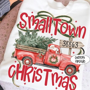 May include: White sweatshirt with a red vintage truck carrying a Christmas tree. The text "Small Town Christmas" is displayed in red, with a Santa Claus graphic and a custom zipcode option.