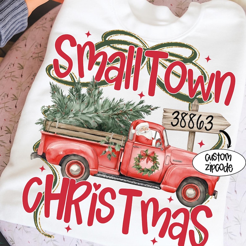May include: White sweatshirt with a red vintage truck carrying a Christmas tree. The text "Small Town Christmas" is displayed in red, with a Santa Claus graphic and a custom zipcode option.