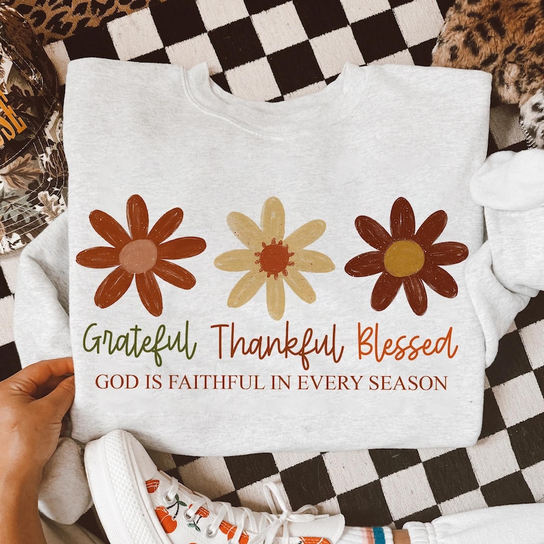 May include: A light gray sweatshirt featuring three stylized flowers in shades of brown and yellow. The words "Grateful Thankful Blessed" are printed below the flowers, with the phrase "GOD IS FAITHFUL IN EVERY SEASON" underneath.