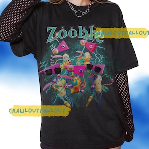 May include: Black t-shirt with a colourful graphic featuring the word "Zooble" in teal above a group of cartoon characters wearing sunglasses. The characters are in front of a teal and green background with lightning bolts.