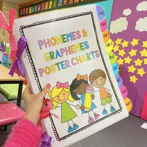 May include: A white binder with a colorful title "PHONEMES & GRAPHEMES POSTER CHARTS". The binder has colorful tabs with letter sounds written on them, such as "a", "ae", "ar", "air", "b", "d", "el", "lee", and "ear".